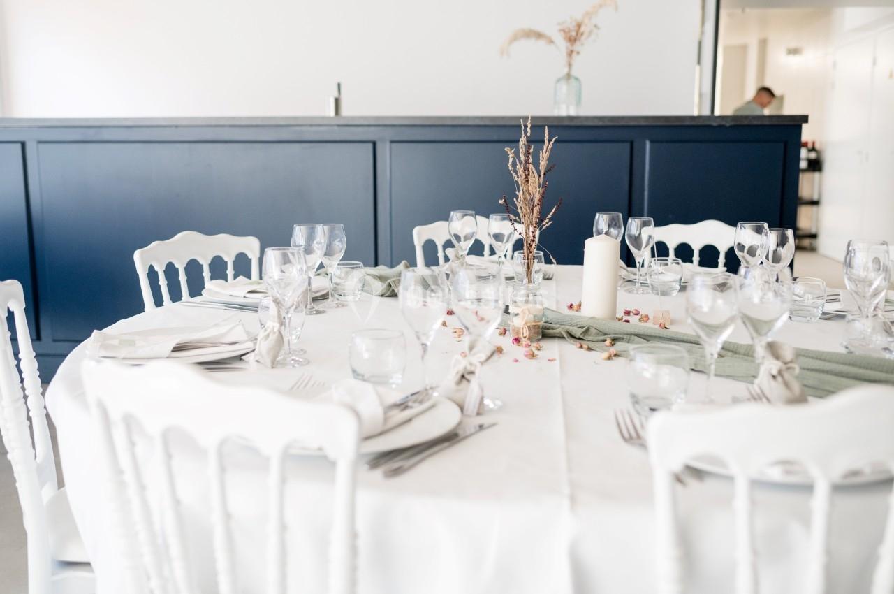 Table ronde dressée avec une nappe blanche, verres, couverts, bougie et décoration florale, devant un comptoir bleu.