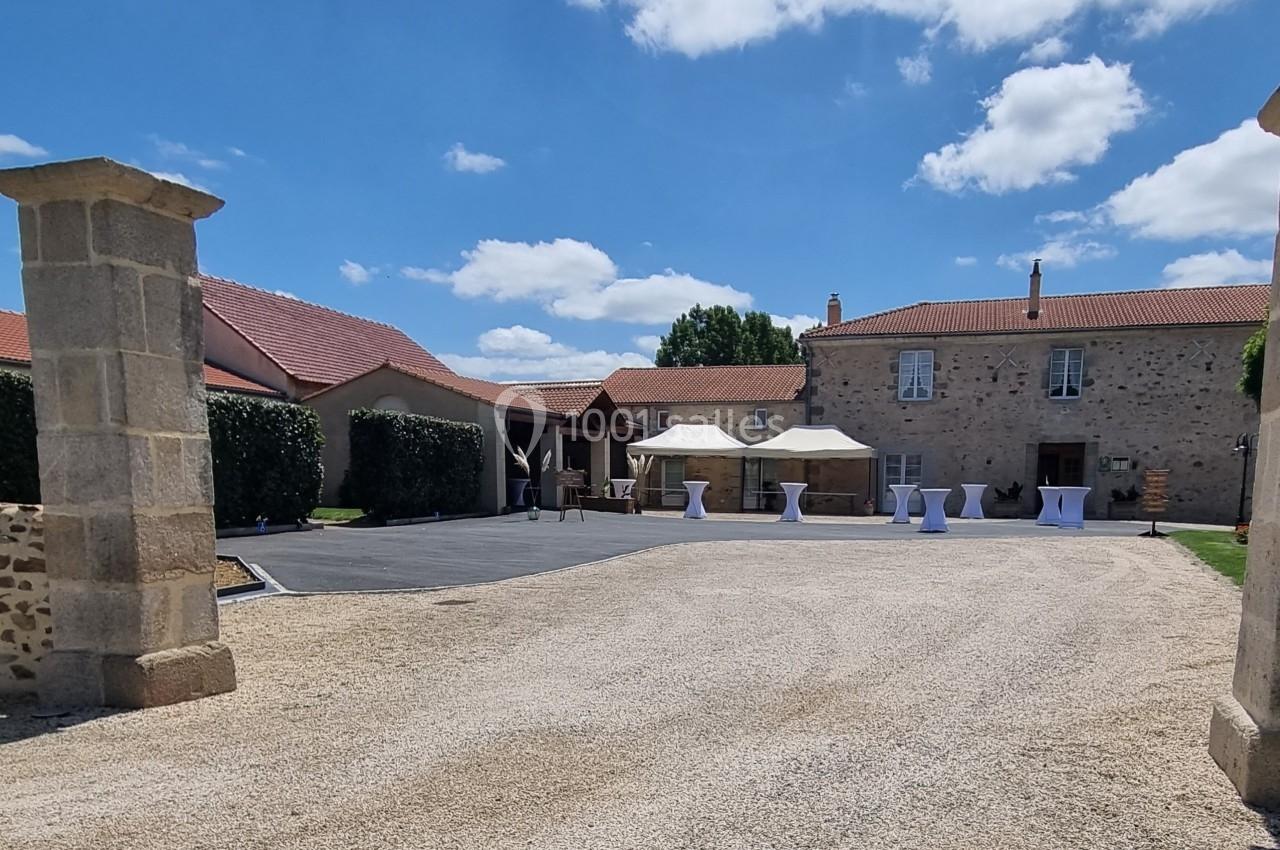 Cour d'une grande bâtisse en pierre avec des tentes blanches, tables hautes et ciel bleu en arrière-plan.