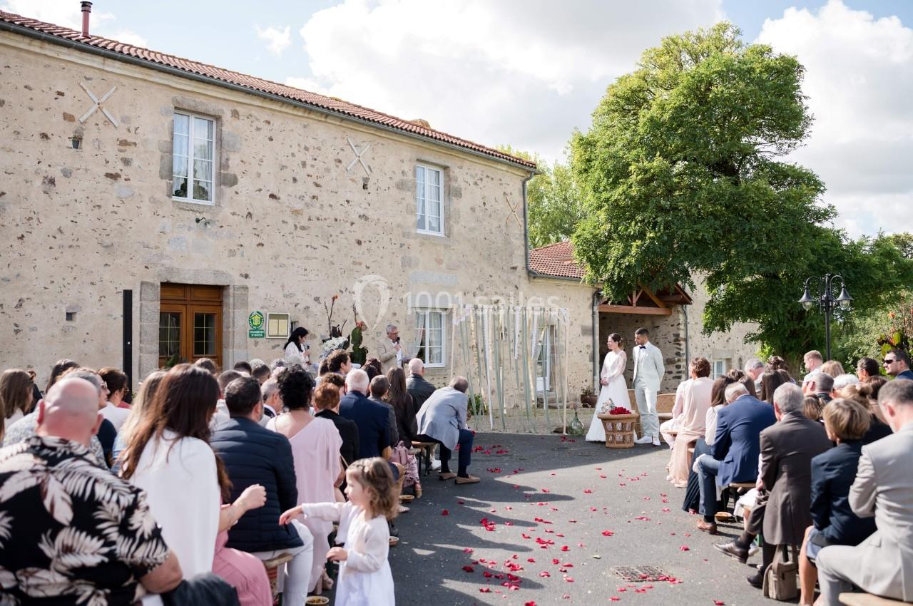 Cérémonie de mariage en plein air devant une bâtisse en pierre, avec invités assis et allée décorée de pétales rouges.