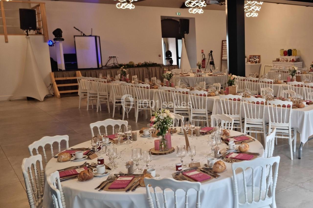 Salle de réception décorée pour un événement, avec tables rondes dressées, fleurs et scène en arrière-plan.