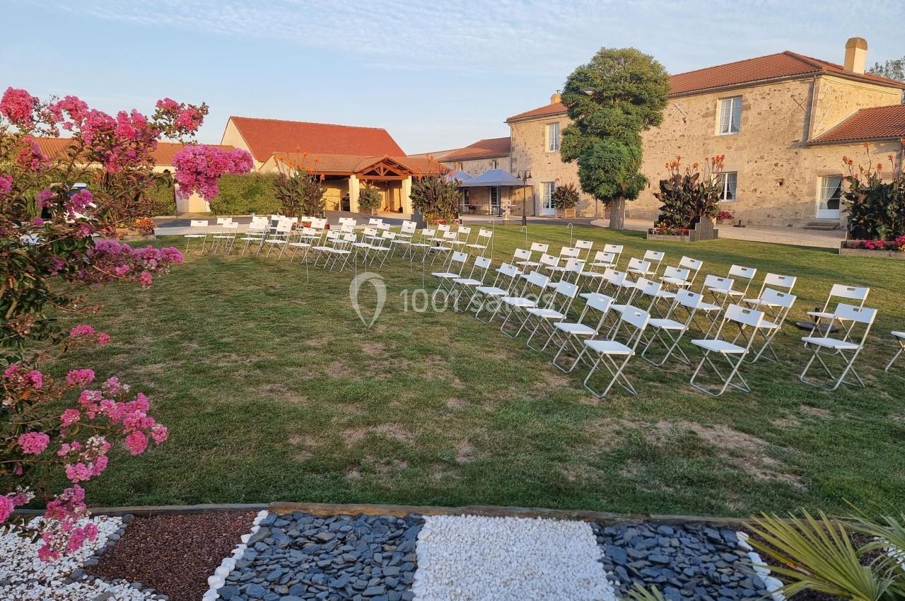 Chaises blanches disposées en extérieur sur une pelouse, entourées de fleurs et bâtiments en pierre sous un ciel dégagé.