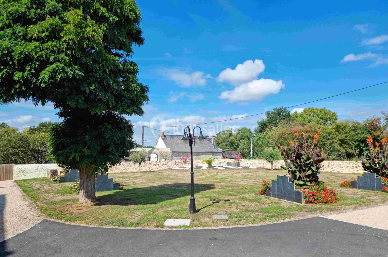 Espace vert avec pelouse, lampadaire central, arbres et fleurs, entouré de murs en pierre et maisons en arrière-plan.