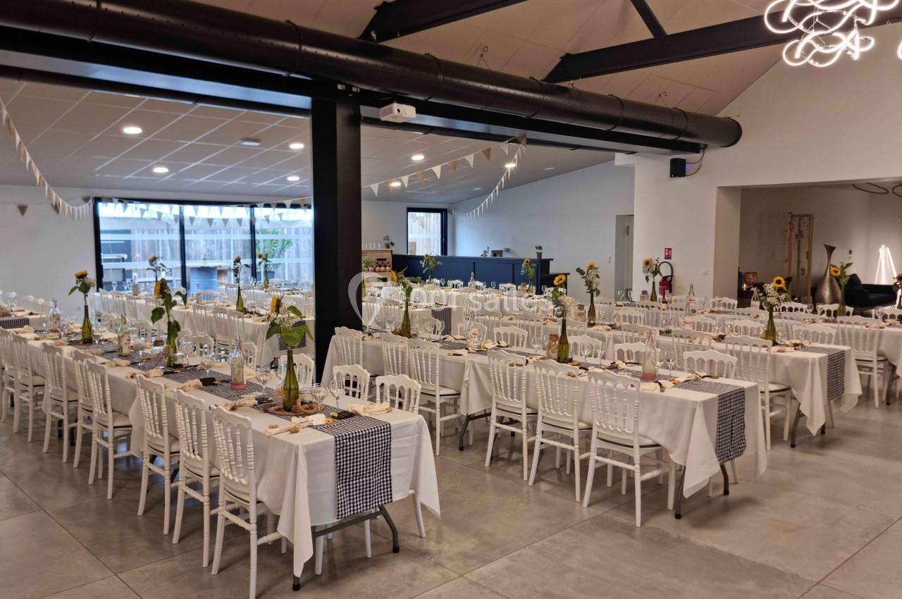 Salle de réception décorée avec des tables alignées, nappes blanches, chaises blanches et centres de table fleuris.