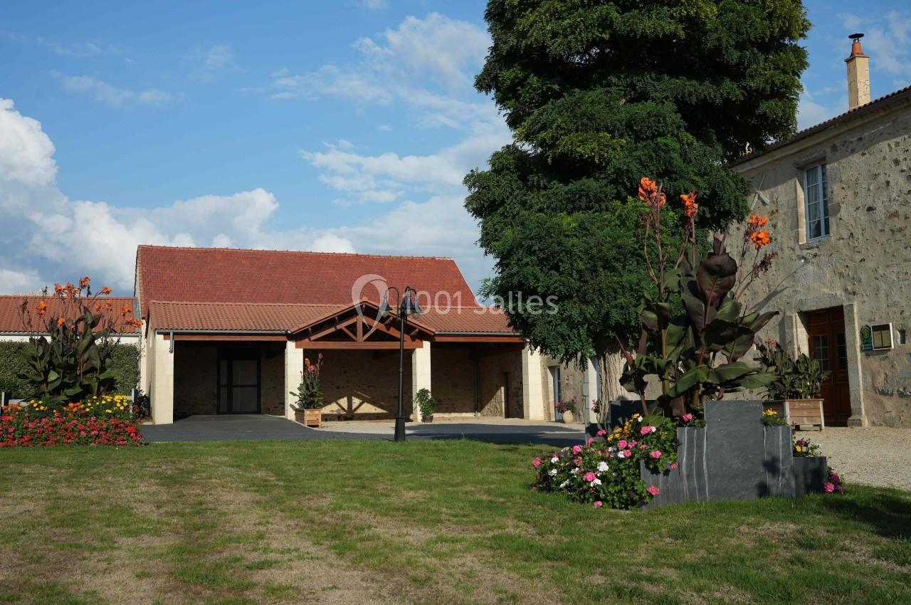 Bâtiment en pierre avec toit rouge, jardin fleuri et grand arbre sous un ciel partiellement nuageux.