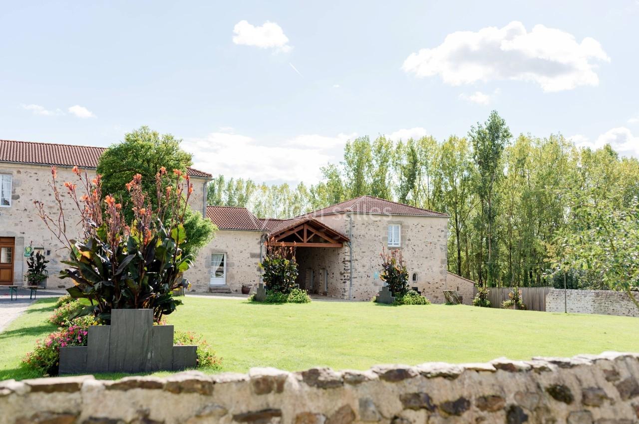 Bâtisse en pierre entourée de verdure, avec un jardin fleuri et un ciel dégagé en arrière-plan.