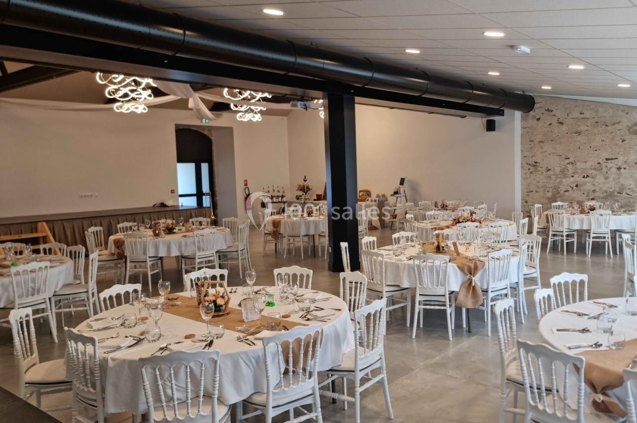 Salle de réception décorée avec des tables rondes dressées, chaises blanches et éclairage suspendu moderne.