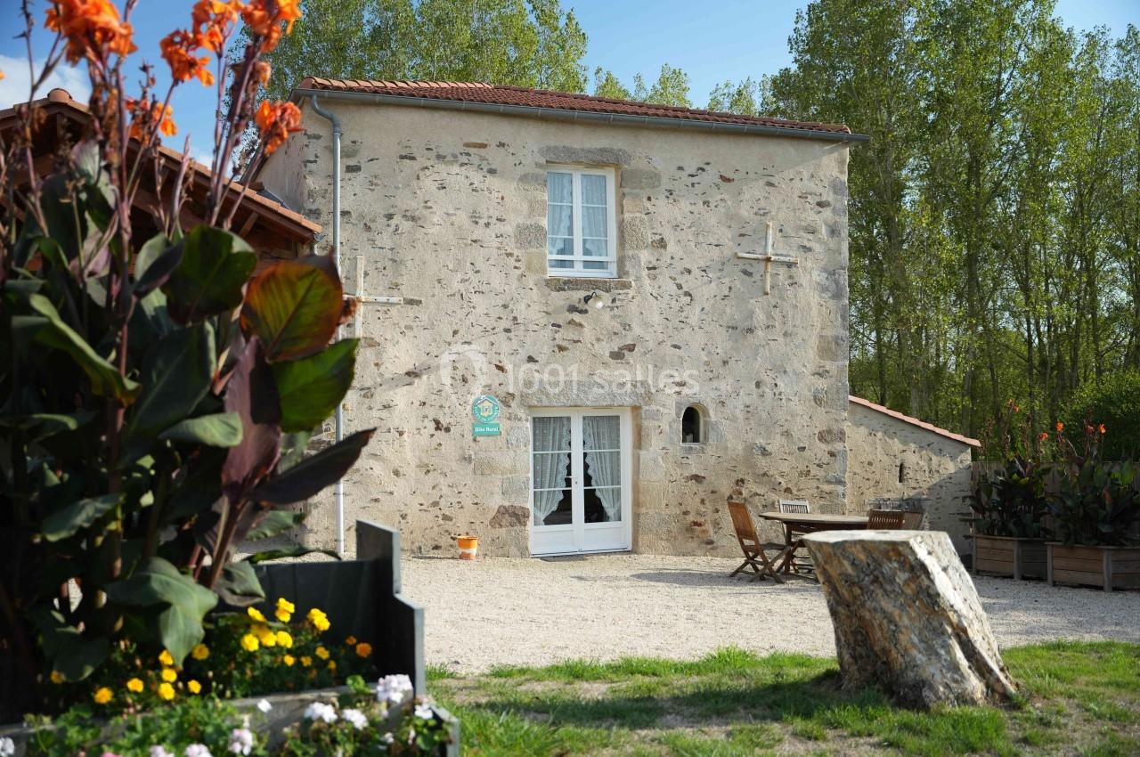 Façade d'une maison en pierre avec porte-fenêtre, mobilier de jardin et fleurs au premier plan, entourée de verdure.
