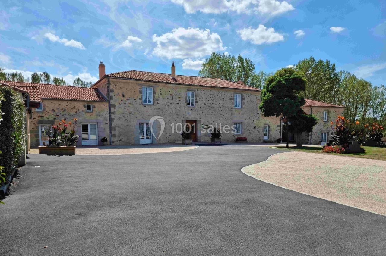 Grande maison en pierre avec toit en tuiles rouges, entourée de végétation et d'une cour asphaltée sous un ciel bleu.