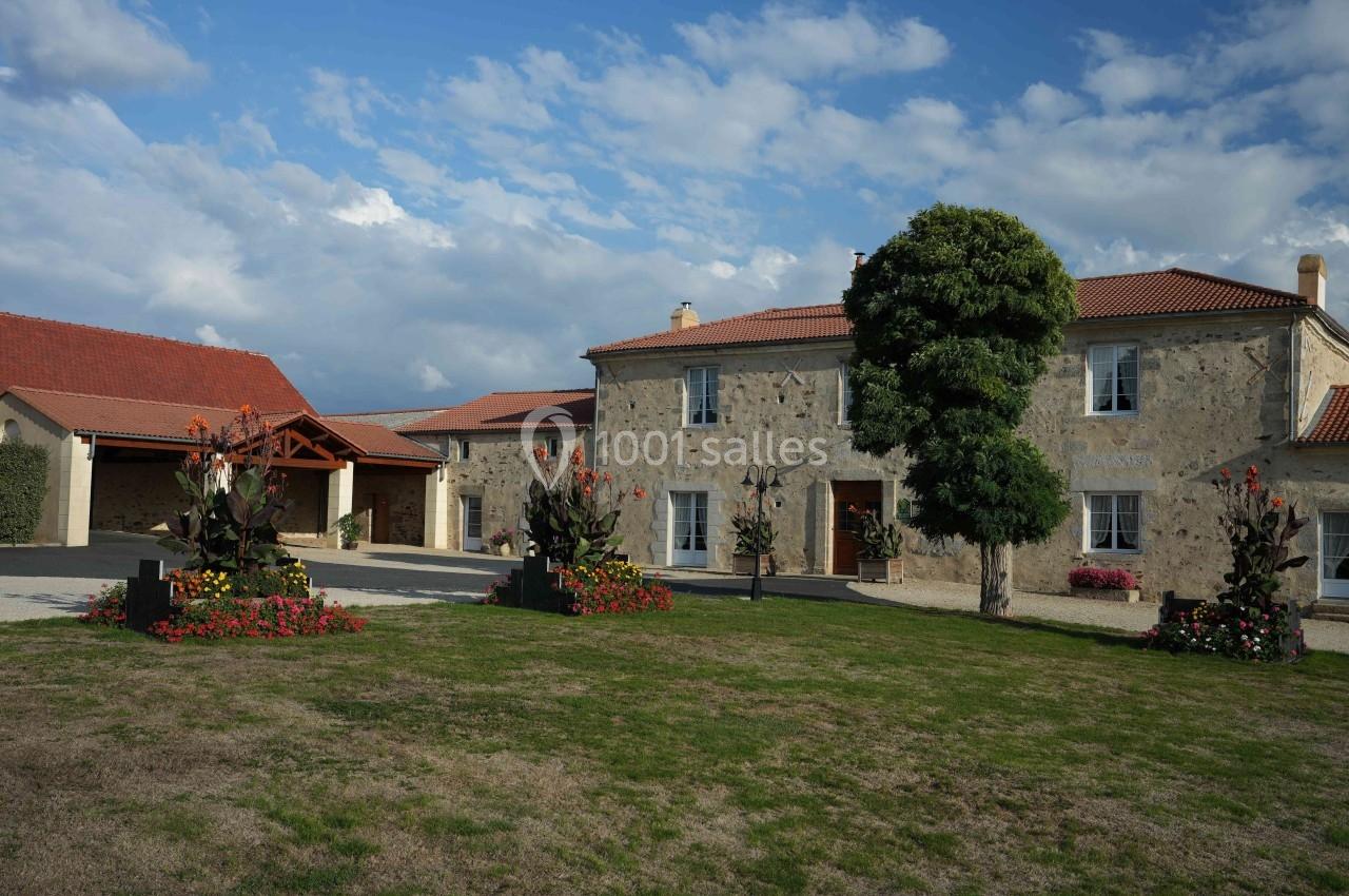 Bâtisse en pierre avec toit en tuiles rouges, entourée de pelouse, fleurs et dépendances sous un ciel partiellement nuageux.