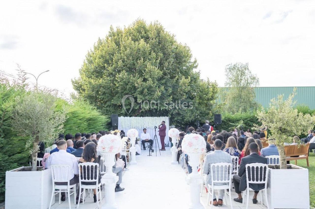 Cérémonie en plein air avec des invités assis face à un officiant devant un décor floral et un grand arbre.