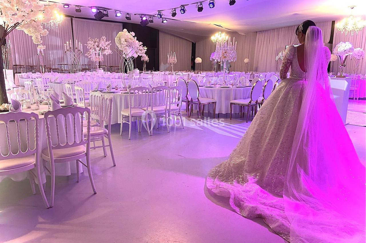 Une mariée en robe blanche observe une salle de réception décorée de fleurs blanches et éclairée de lumières violettes.