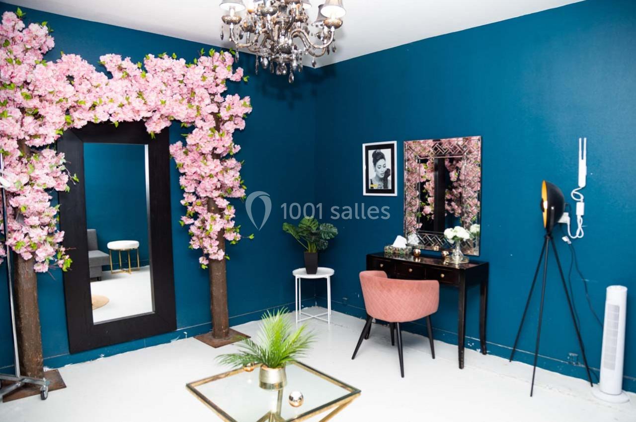 Salle élégante avec murs bleu foncé, miroir encadré de fleurs roses, coiffeuse, fauteuil rose et lustre suspendu.