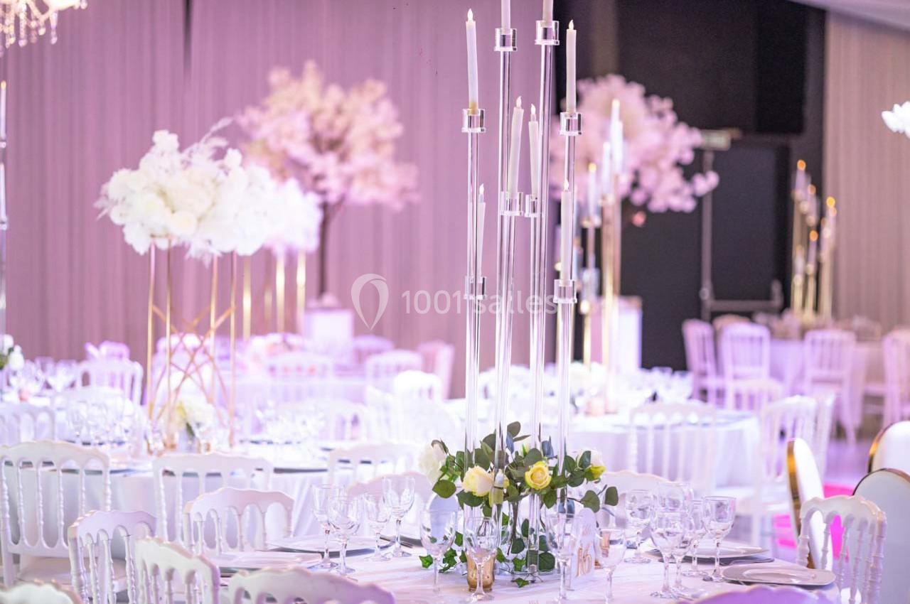 Salle de réception décorée avec des tables dressées, chandeliers modernes et arrangements floraux blancs.