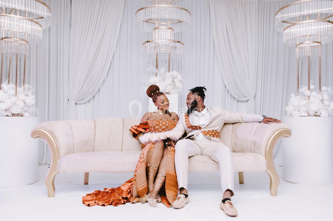 Un couple assis sur un canapé beige dans un décor élégant avec des rideaux blancs et des luminaires suspendus.