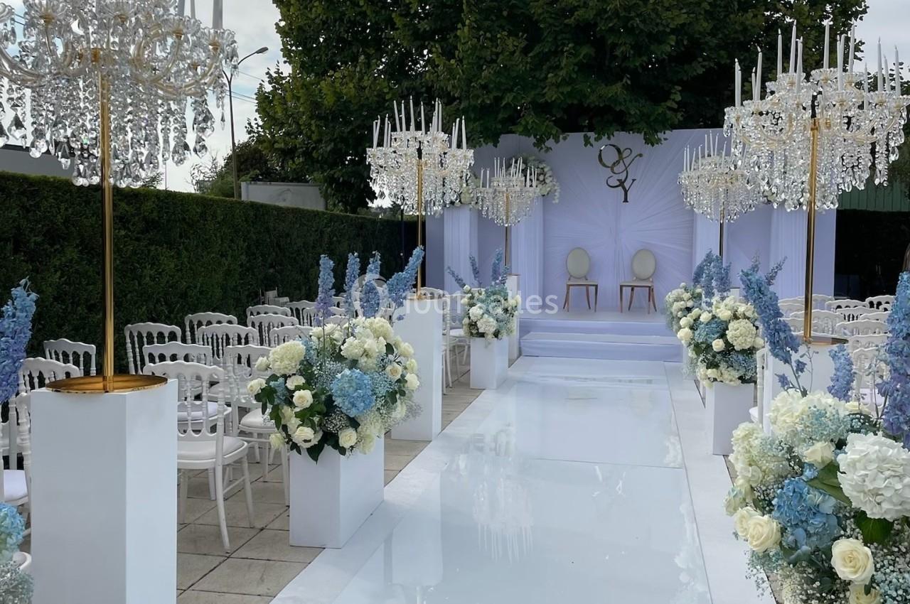 Allée centrale décorée de fleurs blanches et bleues, menant à une scène avec deux chaises, lors d'une cérémonie en extérieur.