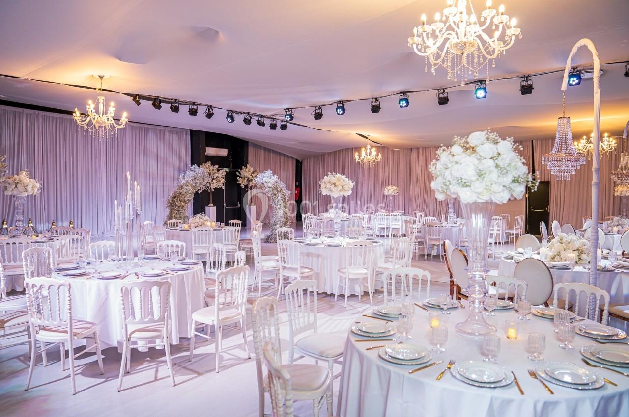 Salle de réception élégante décorée de tables rondes avec nappes blanches, chandeliers et arrangements floraux.