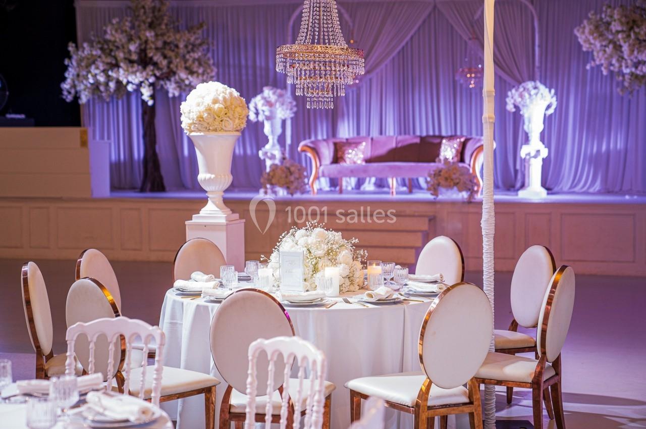 Salle de réception décorée avec des tables rondes élégantes, des chaises blanches et un éclairage violet.