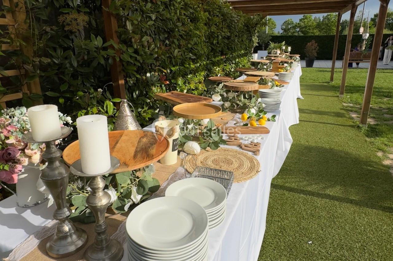 Buffet en extérieur avec nappes blanches, vaisselle, bougies et décorations végétales sur une pelouse.