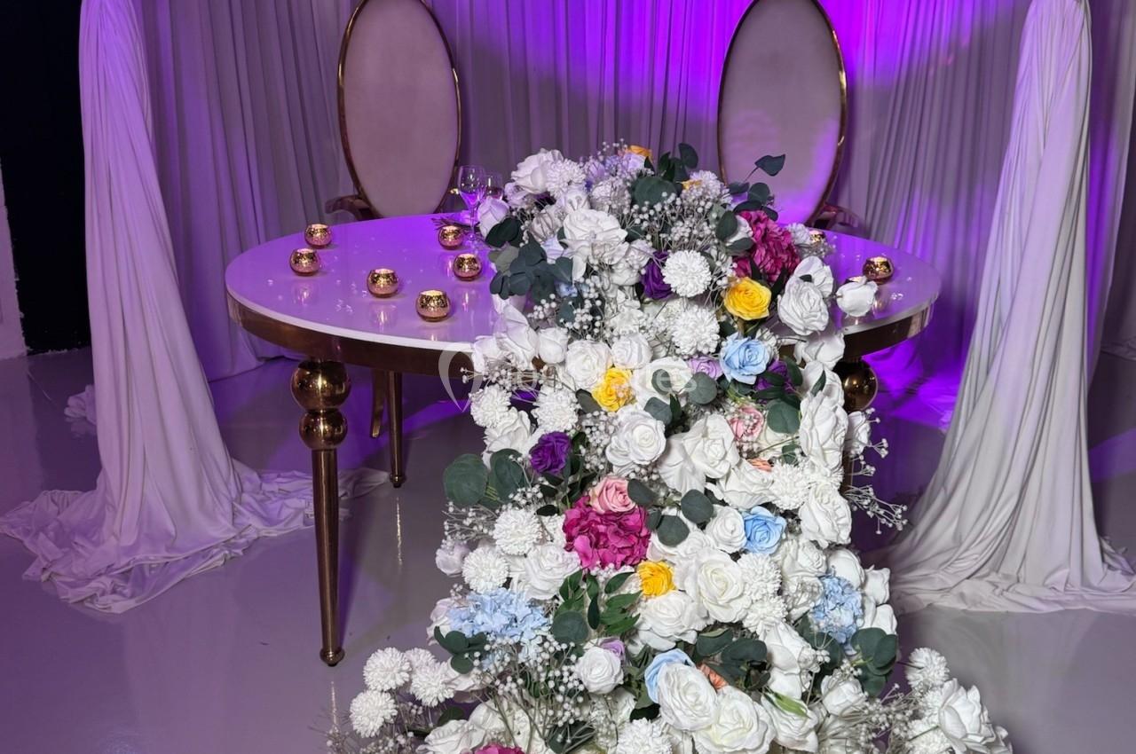 Table décorée avec des chaises élégantes, drapés blancs, éclairage violet et cascade de fleurs multicolores.