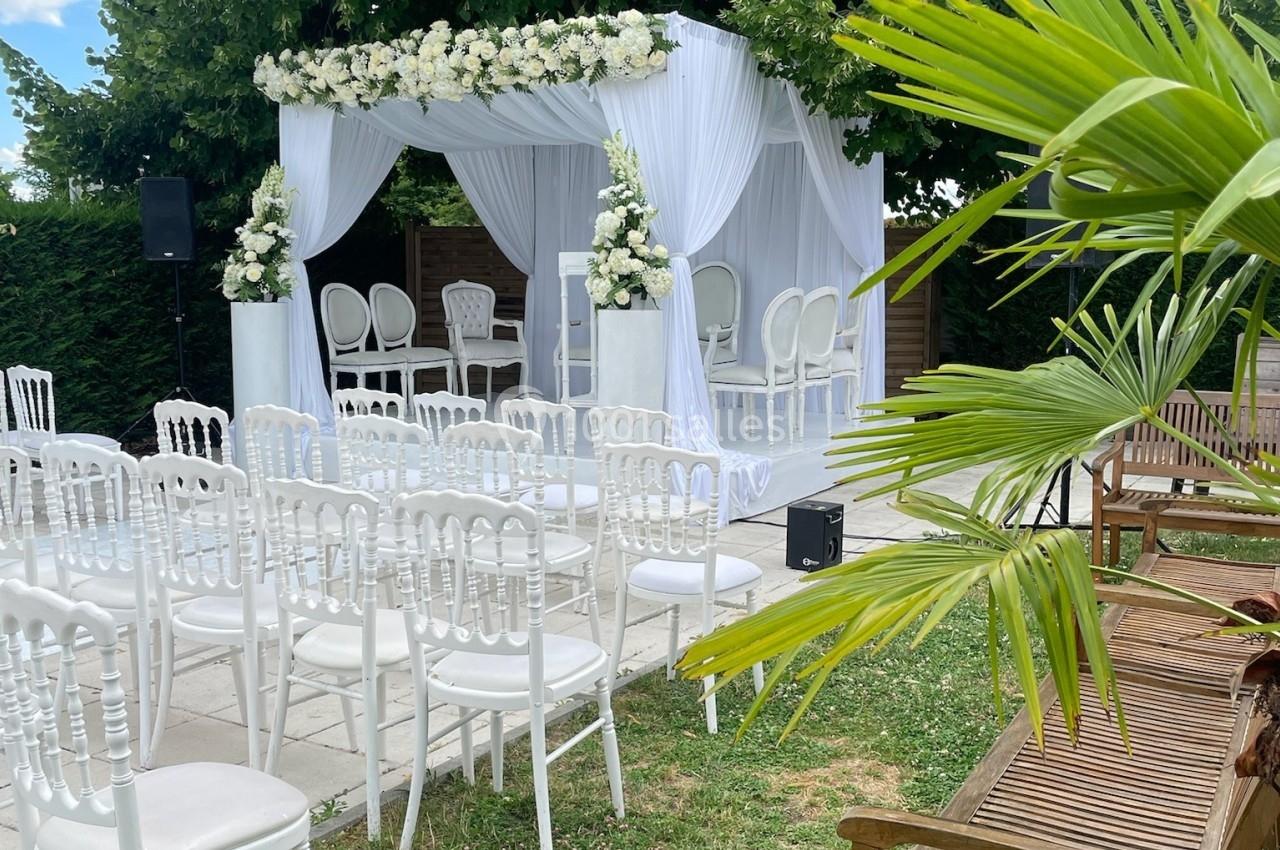 Cérémonie en plein air avec arche fleurie blanche, chaises blanches alignées et bancs en bois dans un jardin verdoyant.