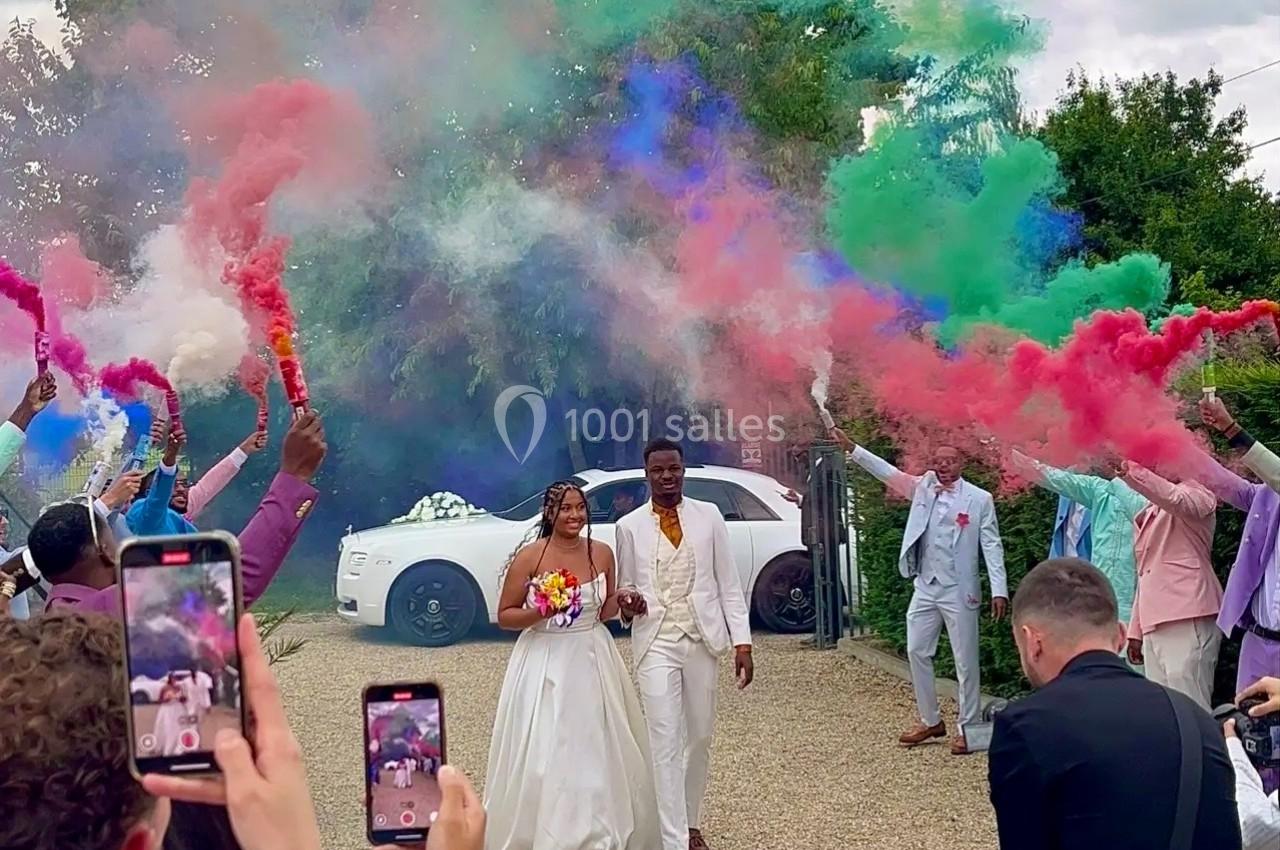 Un couple en tenue de mariage marche sous des fumigènes colorés, entouré d'invités prenant des photos.