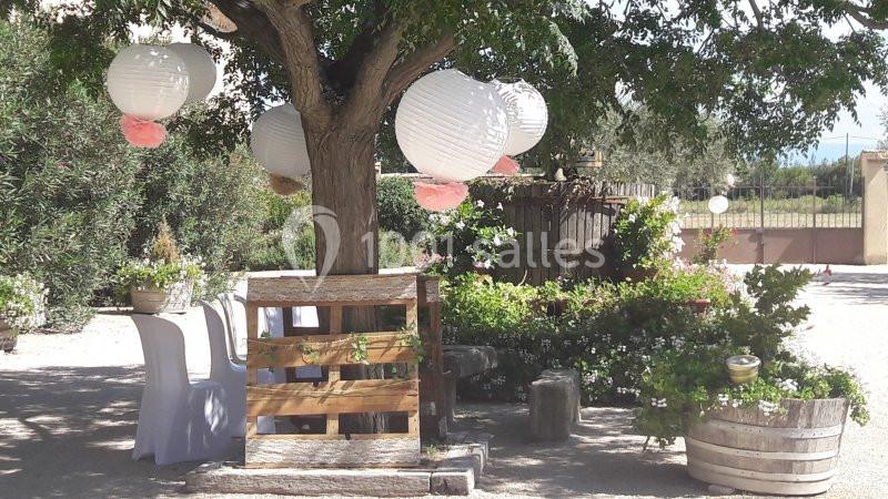 Décoration extérieure avec lanternes blanches suspendues à un arbre, table en bois et plantes en pots.