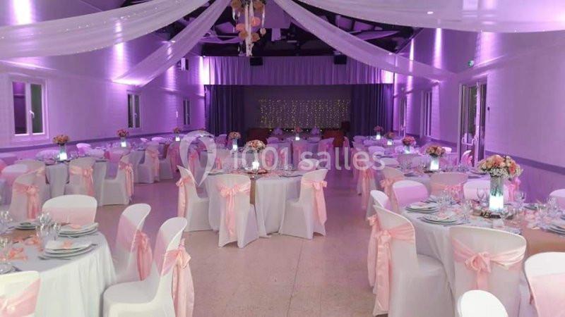 Salle de réception décorée avec des nappes blanches, des chaises ornées de rubans roses et un éclairage violet.