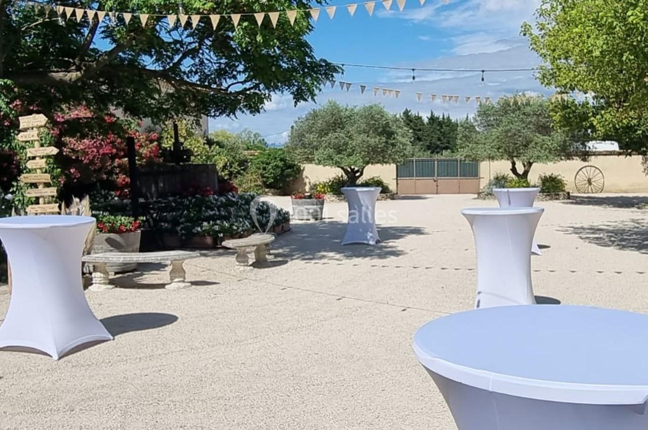 Cour extérieure aménagée avec des tables hautes drapées de blanc, des arbres, des guirlandes et des fleurs en arrière-plan.