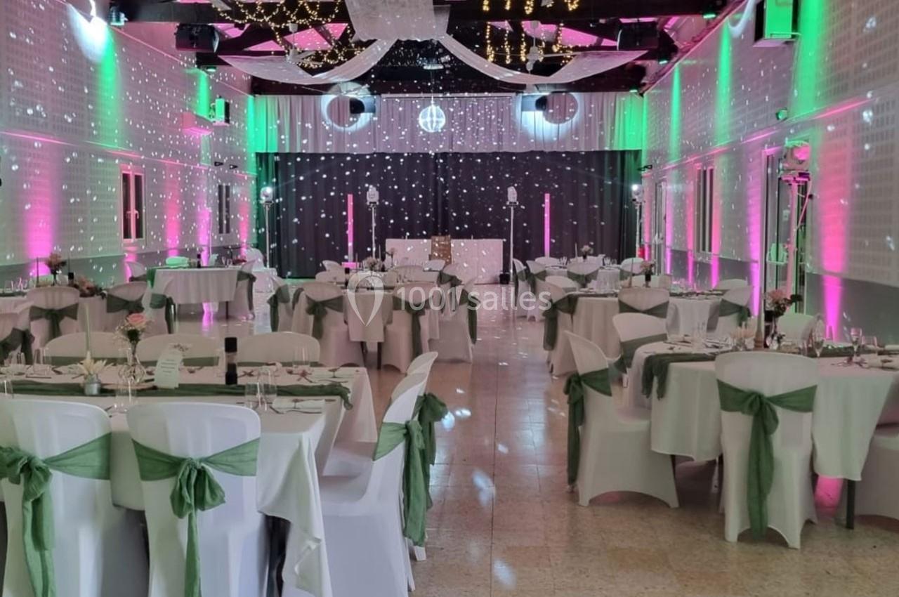 Salle de réception décorée avec des nappes blanches, des rubans verts et un éclairage rose et vert.