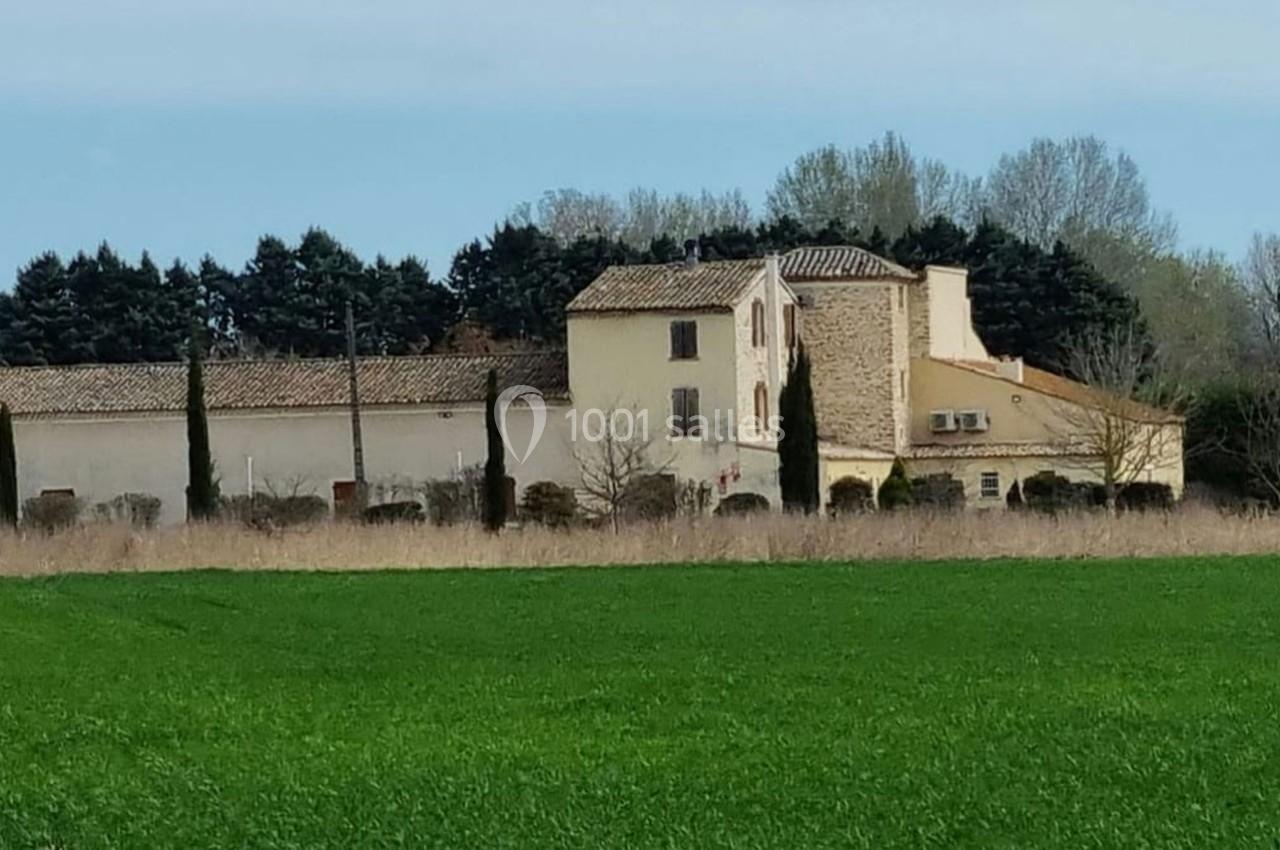 Bâtisse provençale entourée de cyprès et de champs verts, avec des arbres en arrière-plan sous un ciel clair.