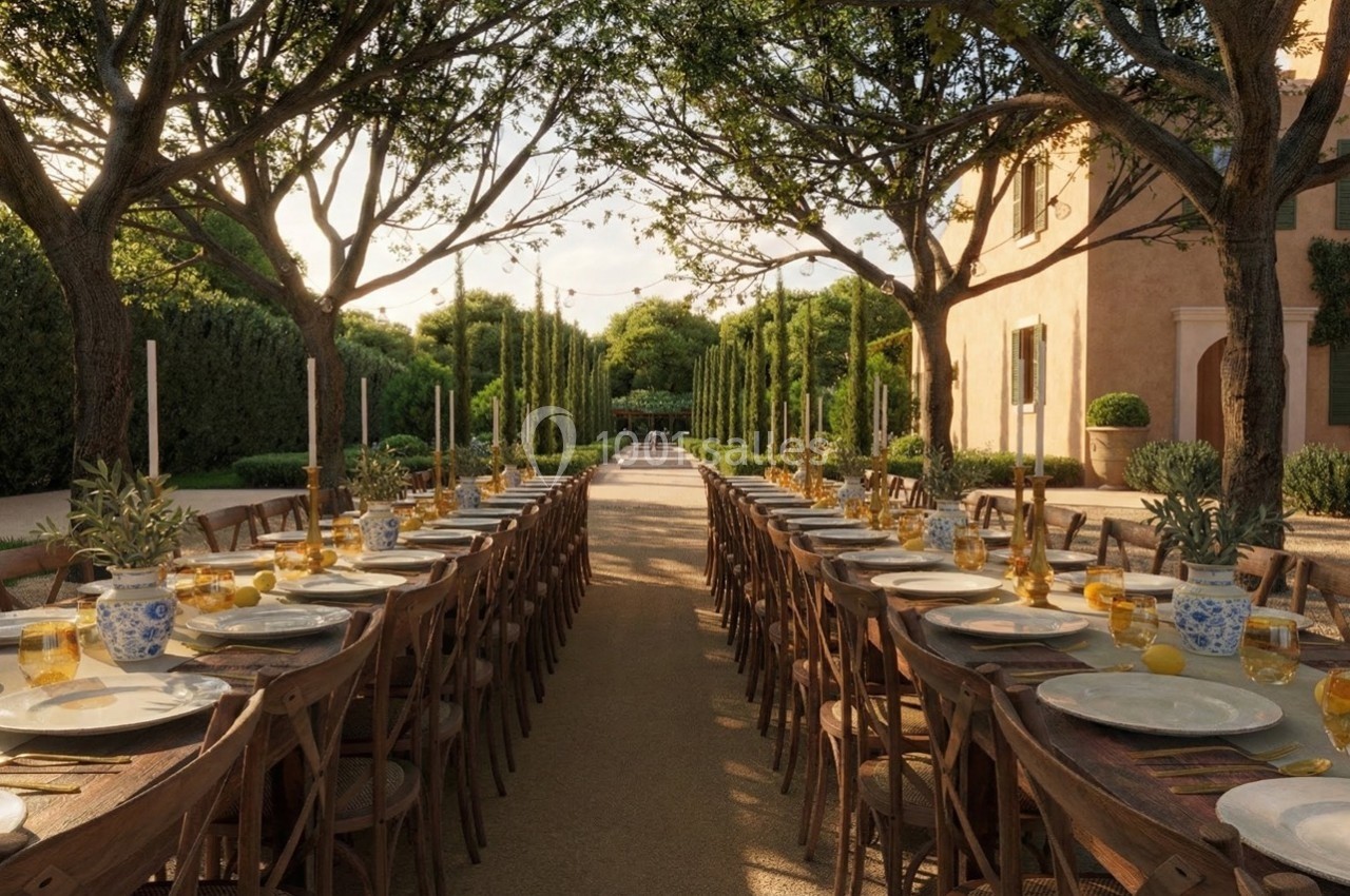 Grande table extérieure dressée avec vaisselle et verres, entourée d'arbres et située devant une maison provençale.