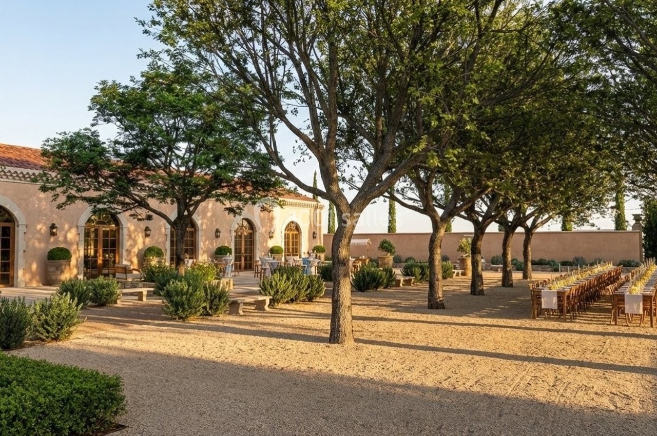 Allée bordée d'arbres menant à une bâtisse en pierre avec tables dressées pour un repas en extérieur.