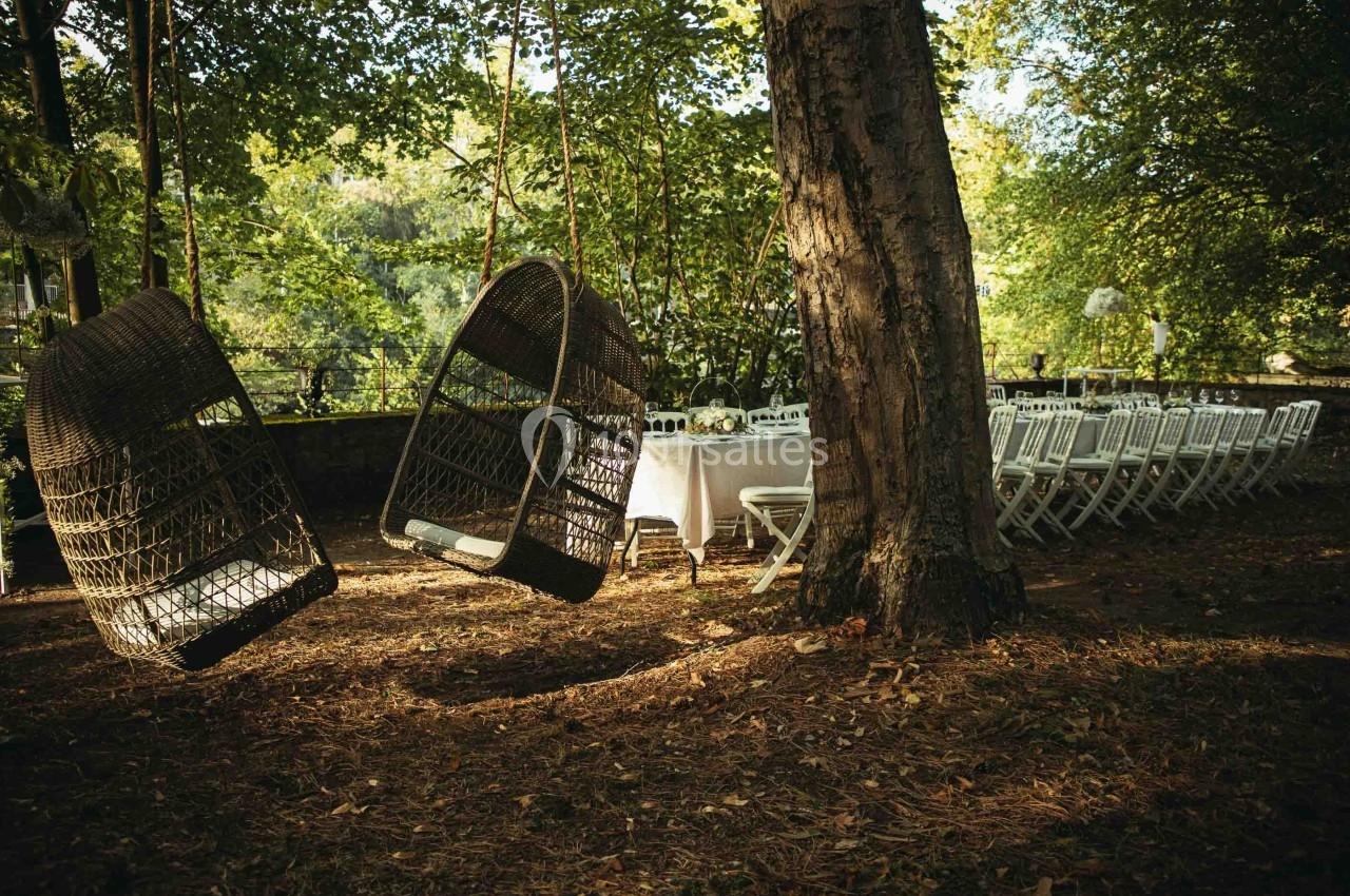 Deux fauteuils suspendus en osier près d'une table dressée avec des chaises blanches dans un jardin ombragé.