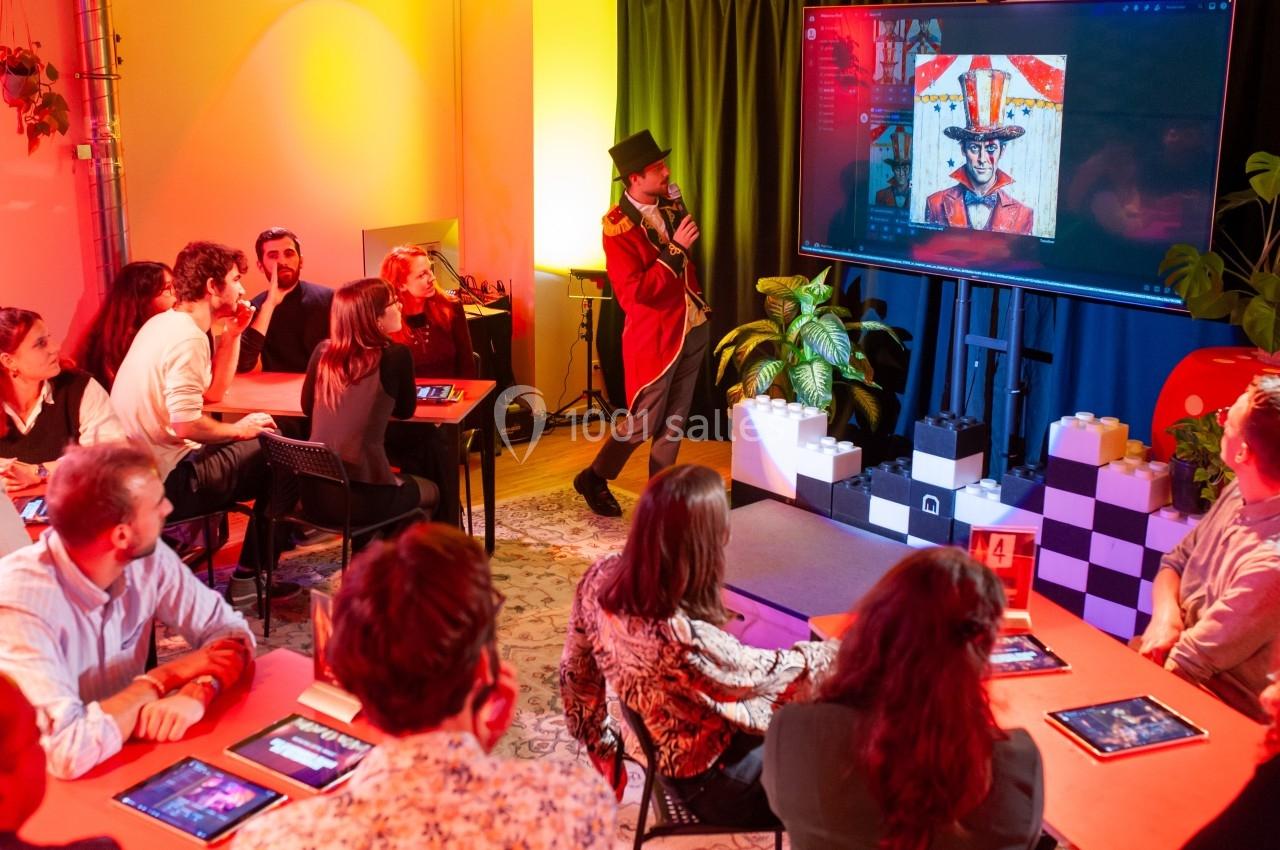 Un animateur en costume présente une image sur un écran à un groupe de personnes assises dans une salle colorée.