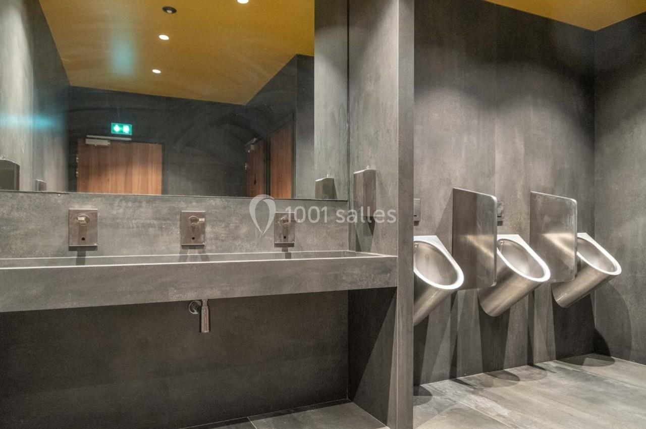 Salle de bain moderne avec lavabo en acier, grand miroir et trois urinoirs alignés sur un mur gris.