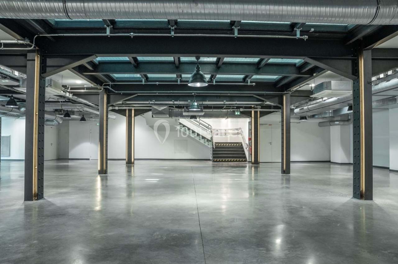 Espace industriel vide avec sol en béton, poutres métalliques apparentes et escalier au fond sous un éclairage artificiel.