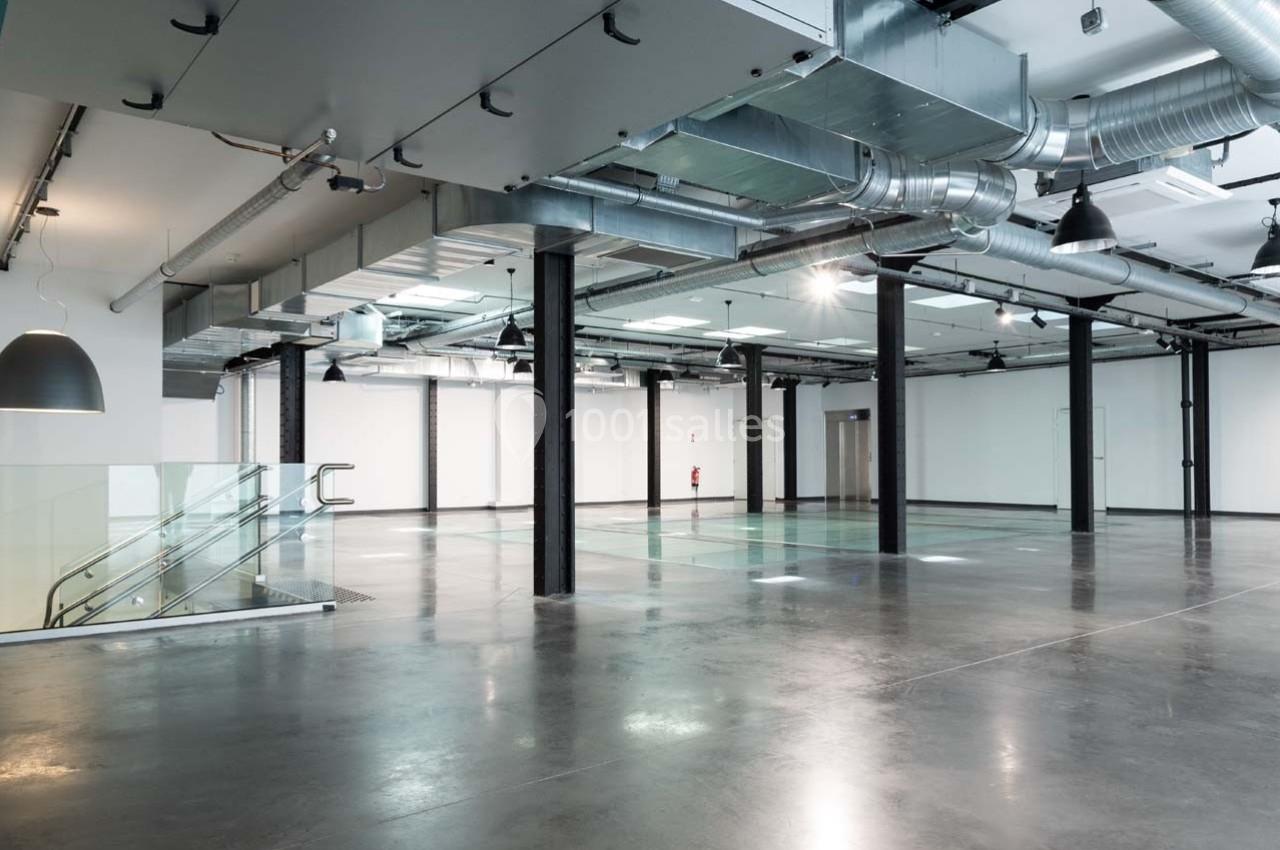 Intérieur d'un espace industriel moderne avec sol en béton poli, escaliers en verre et conduits apparents au plafond.