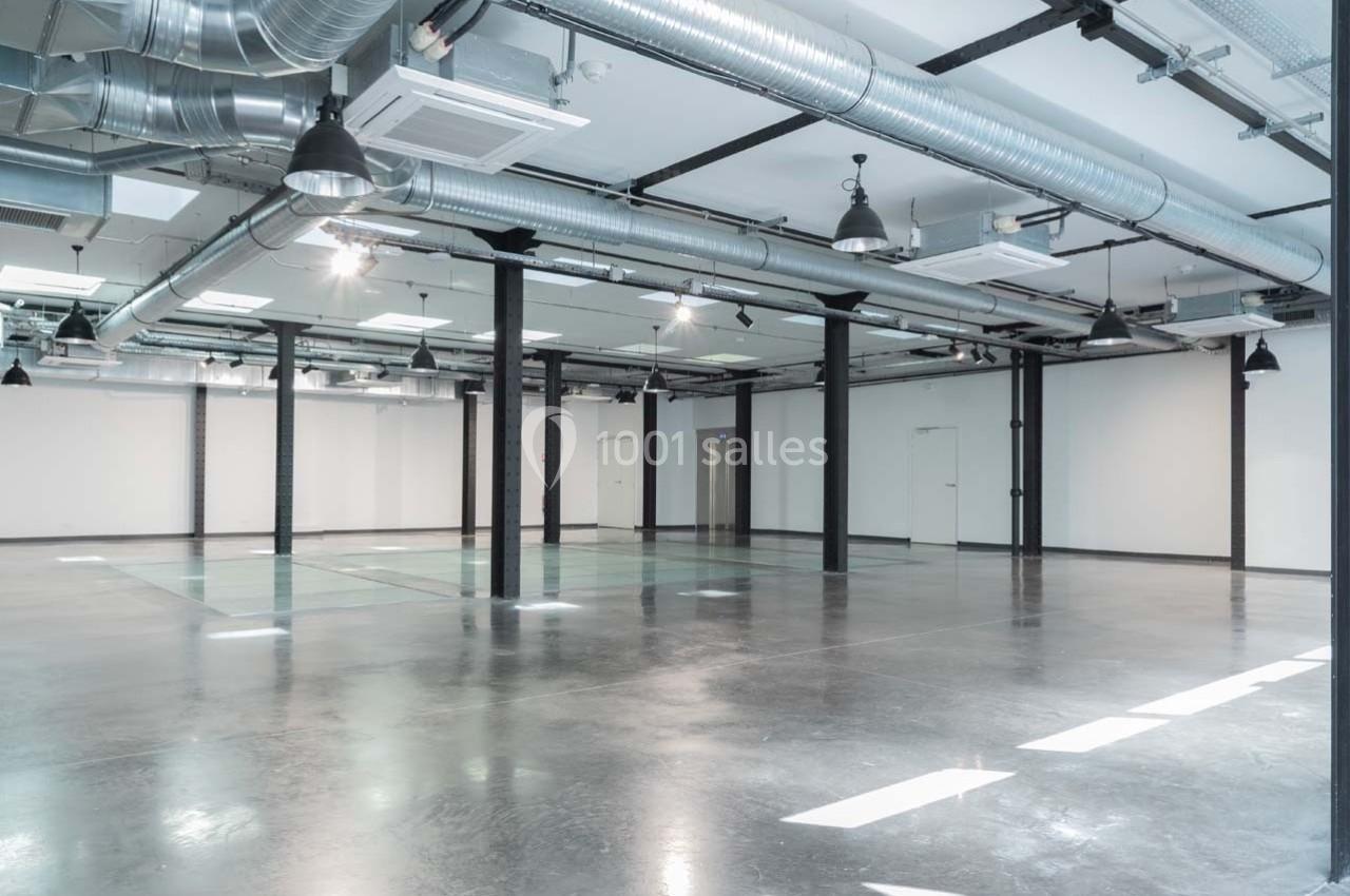 Intérieur d'un espace industriel vide avec sol en béton, murs blancs, poutres métalliques et conduits apparents.