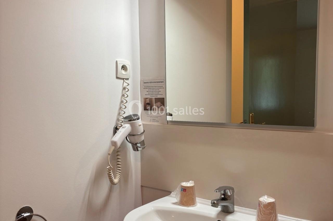 Lavabo avec miroir, sèche-cheveux mural et deux gobelets en plastique posés sur le rebord dans une salle de bain.