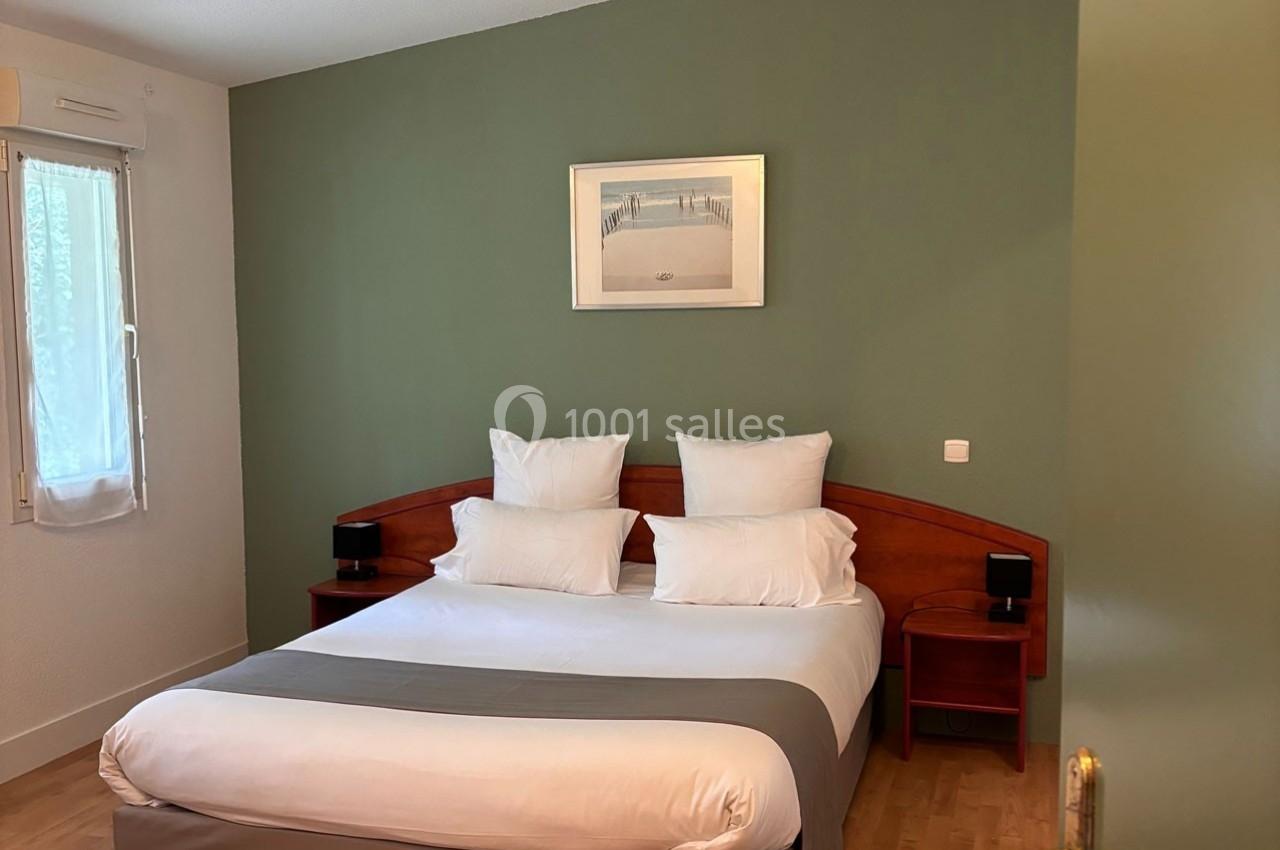 Lit double avec draps blancs, tête de lit en bois, mur vert avec tableau, fenêtre et rideau blanc à gauche.