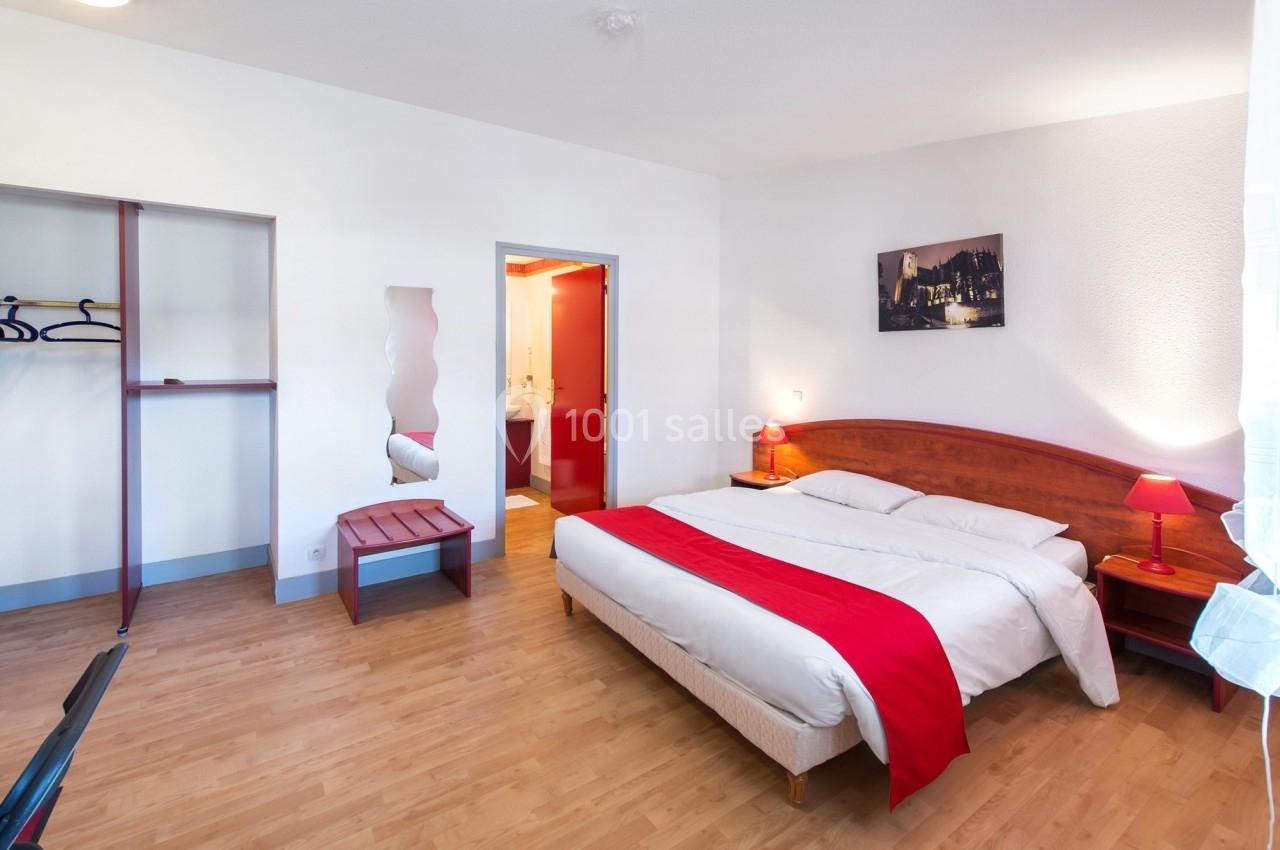 Chambre lumineuse avec lit double, tête de lit en bois, lampe rouge, armoire ouverte et salle de bain attenante.