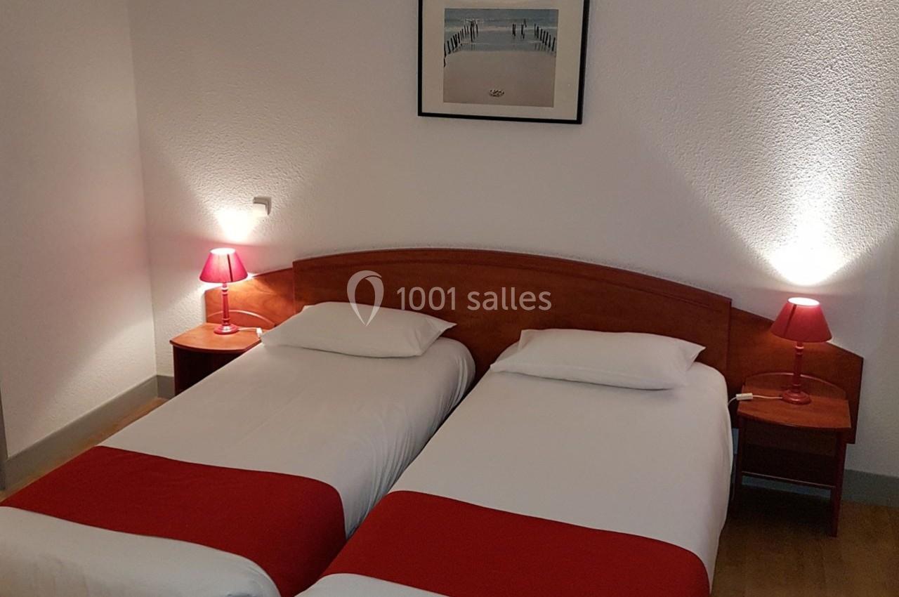 Deux lits simples avec draps blancs et couvre-lits rouges, encadrés par des lampes de chevet rouges, dans une chambre sobre.