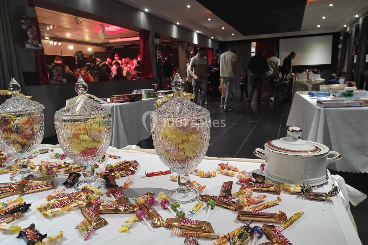 Bonbons et friandises disposés sur une table blanche, avec des bocaux en verre, dans une salle animée en arrière-plan.