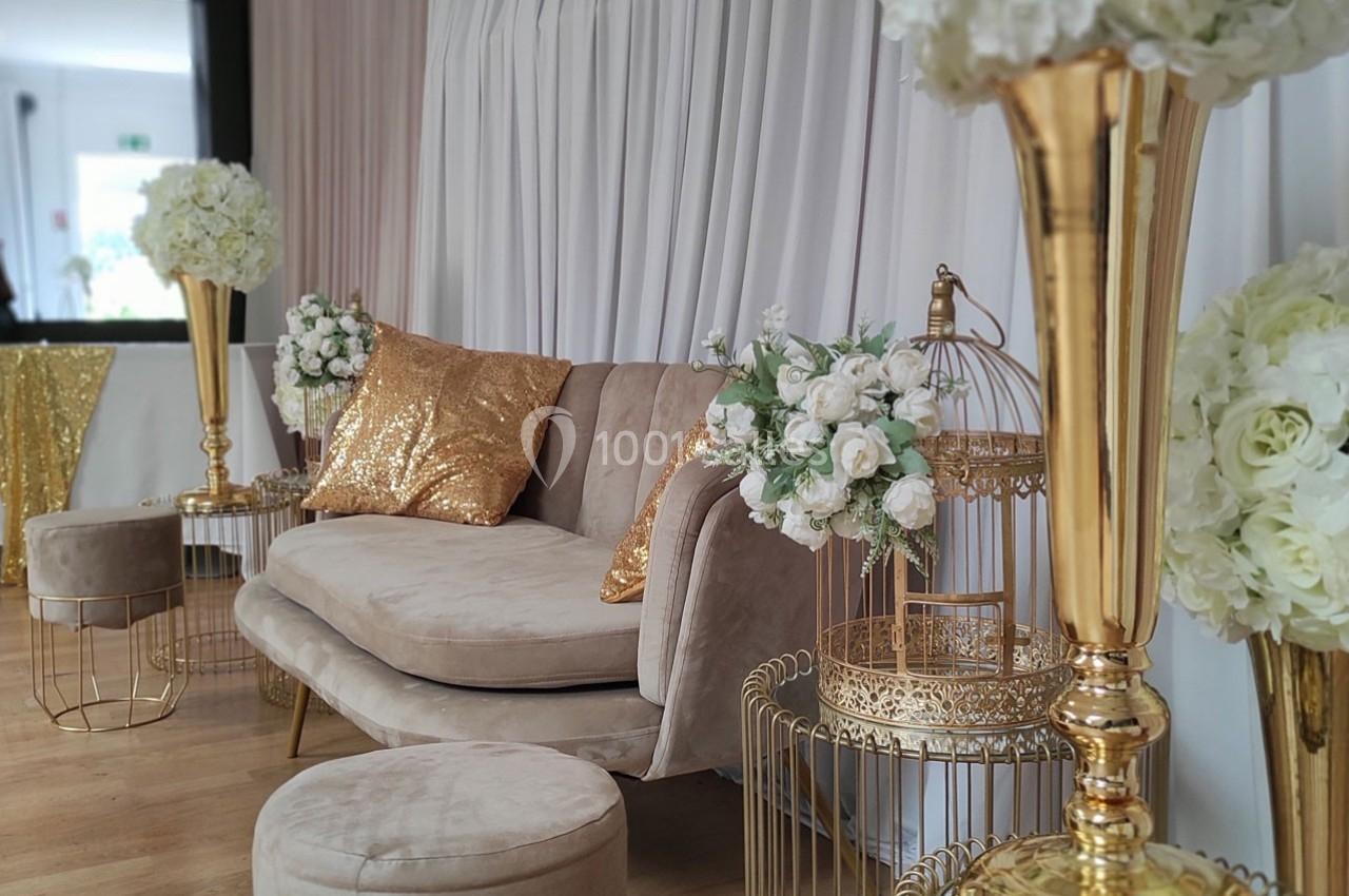 Canapé beige avec coussins dorés, entouré de décorations florales blanches et d'éléments dorés sur un sol en bois.