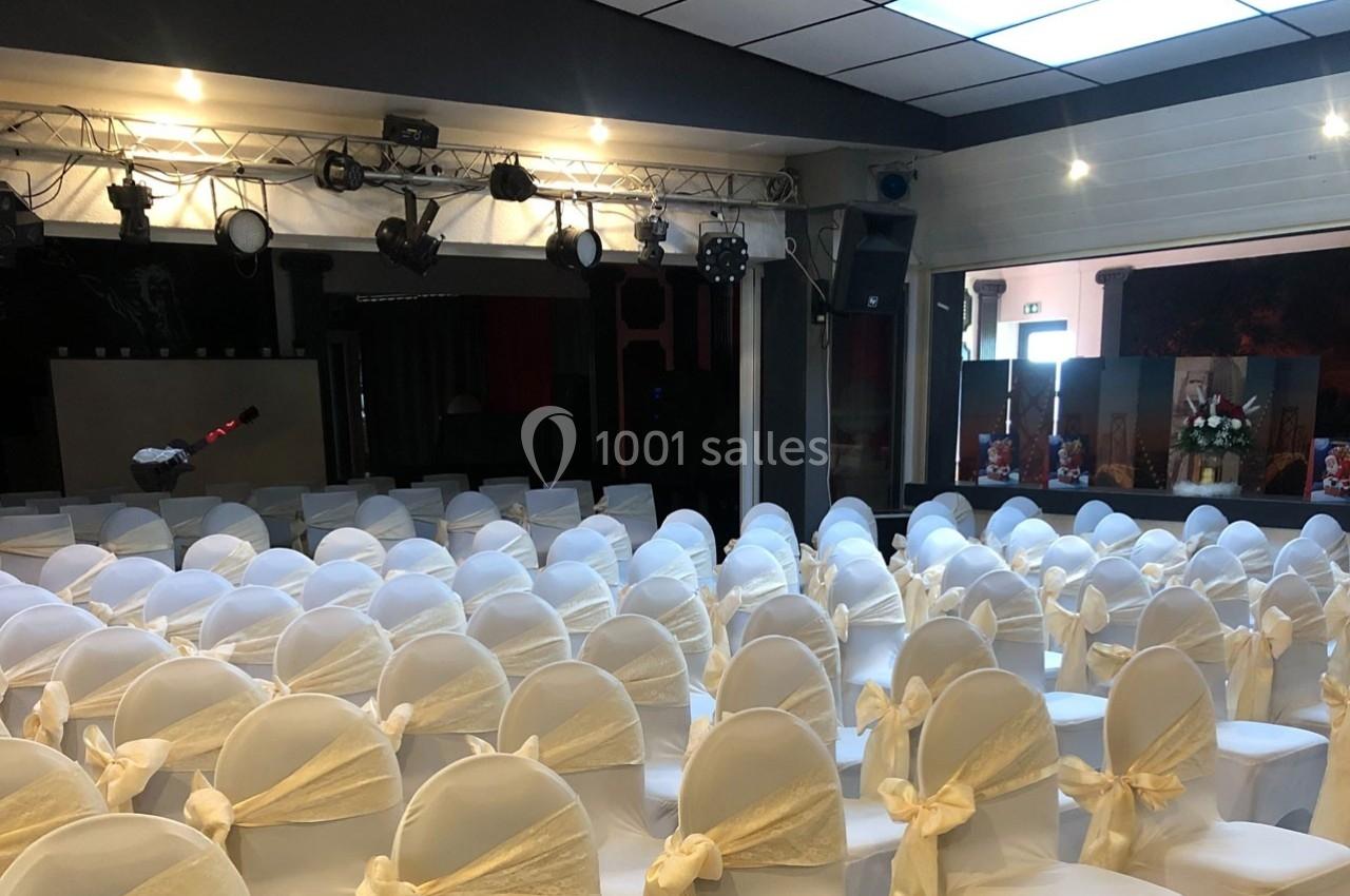 Salle de réception avec des chaises blanches décorées de nœuds, disposées face à une scène équipée de projecteurs.