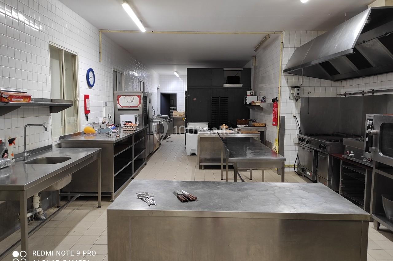 Cuisine professionnelle équipée avec plans de travail en inox, fours, plaques de cuisson et carrelage mural blanc.