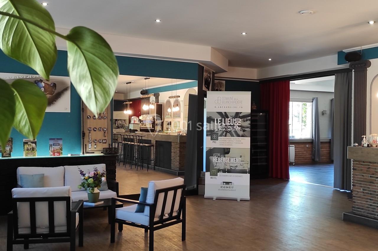 Espace intérieur lumineux avec coin salon, bar moderne, plantes et affiches promotionnelles en arrière-plan.