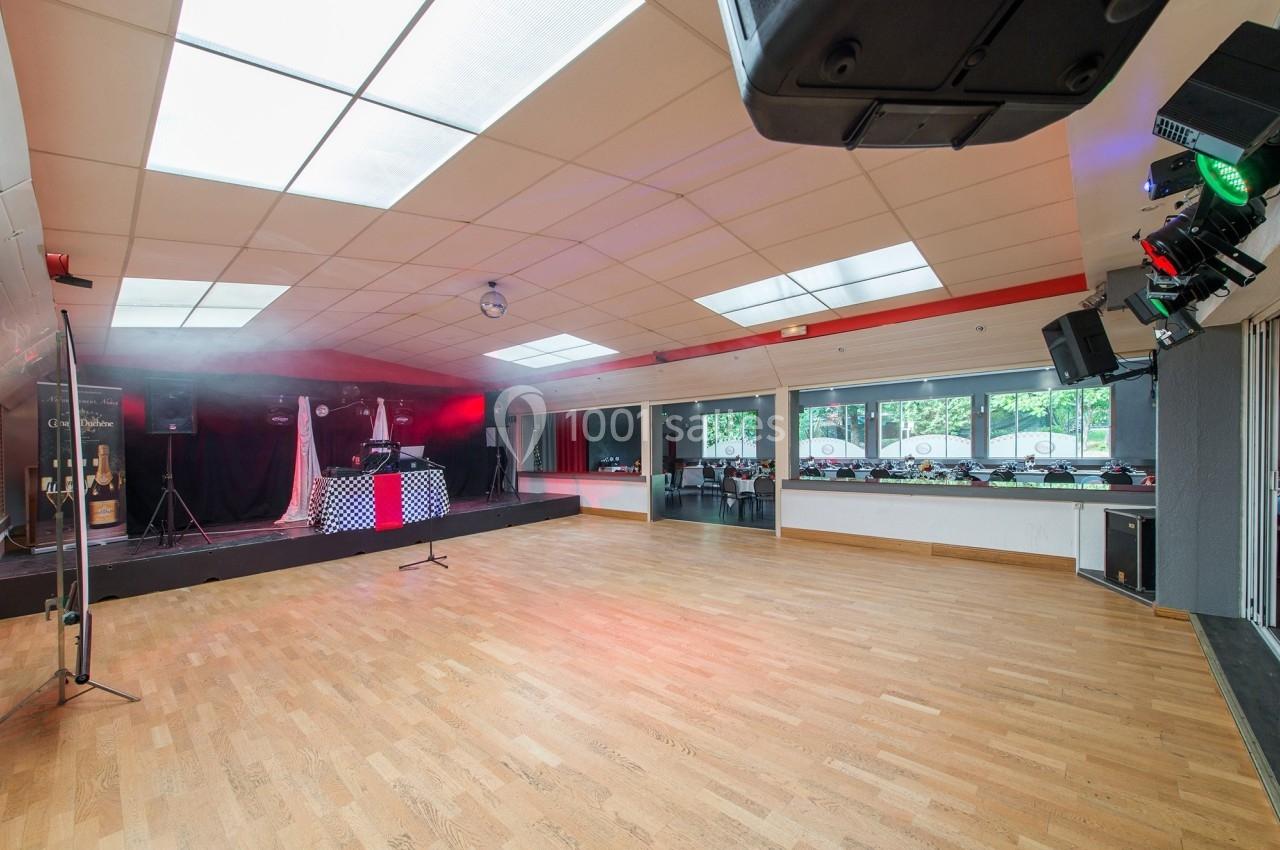 Salle spacieuse avec parquet, scène équipée de matériel audio et éclairage, baies vitrées donnant sur l'extérieur.