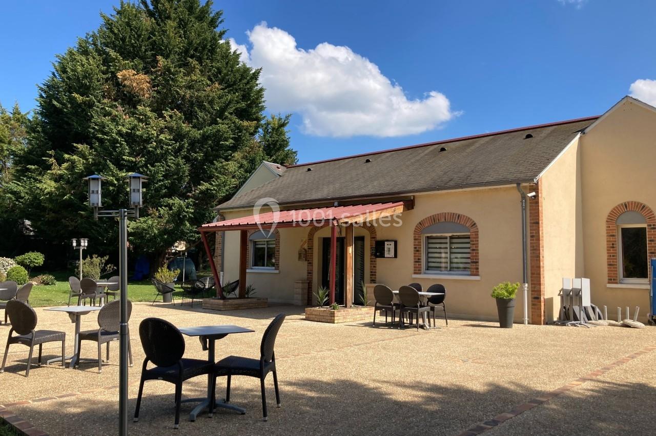Terrasse avec tables et chaises devant un bâtiment beige à toit en ardoise, entouré de végétation et éclairé par le soleil.
