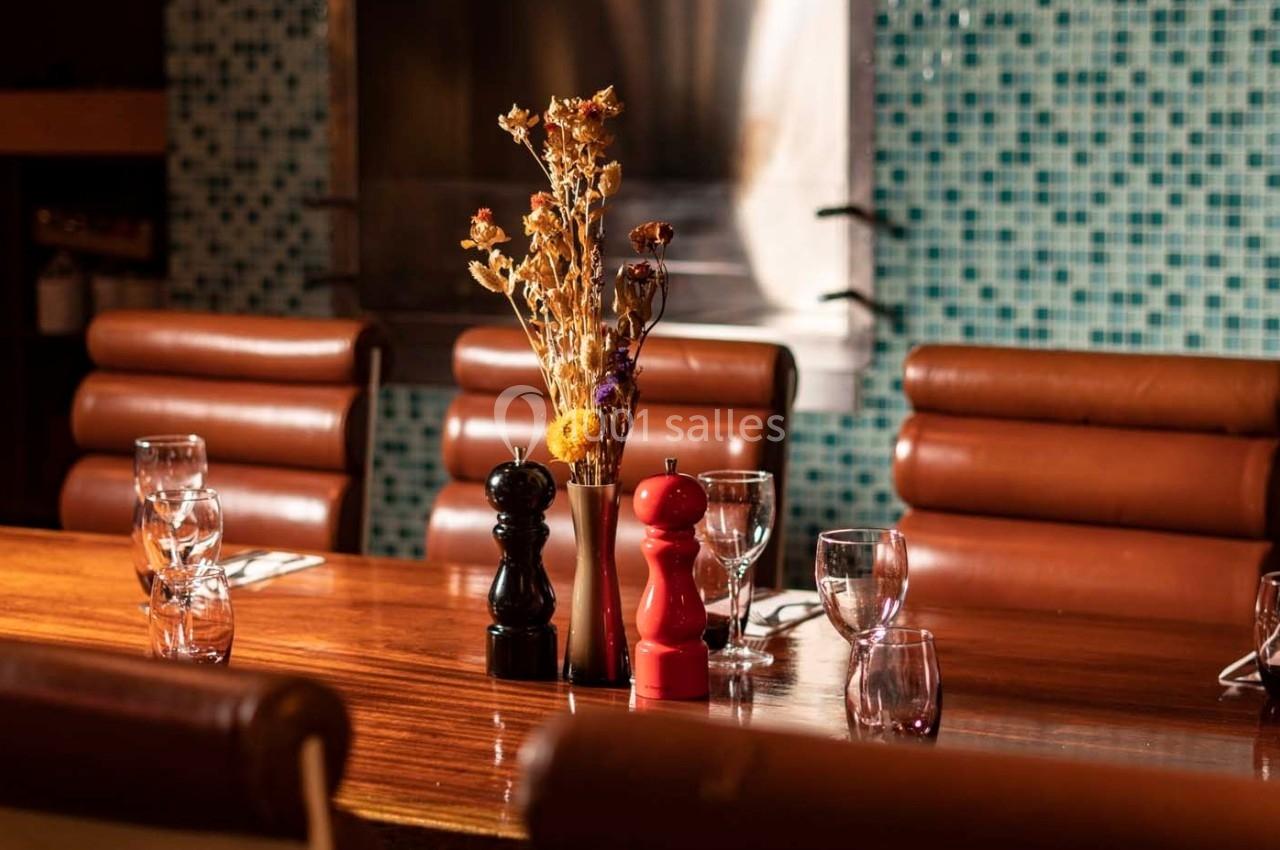 Table en bois avec chaises en cuir, décorée de fleurs séchées, moulins à poivre et verres, devant un mur en mosaïque bleue.